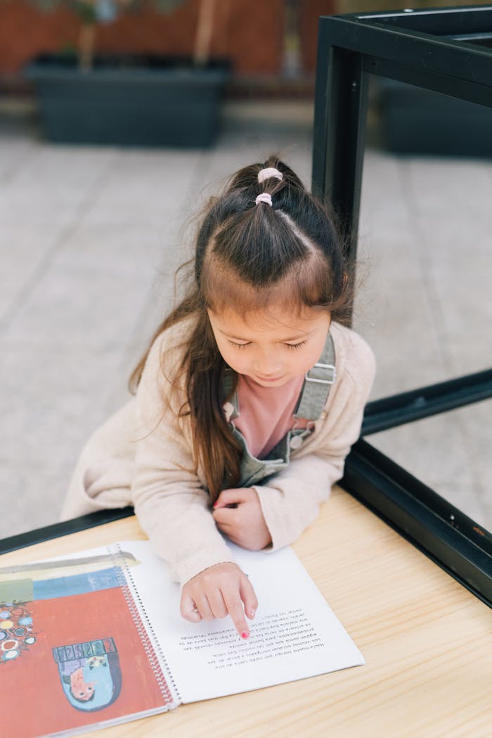 A young girl reading a book outside, focusing on the text with curiosity and concentration.